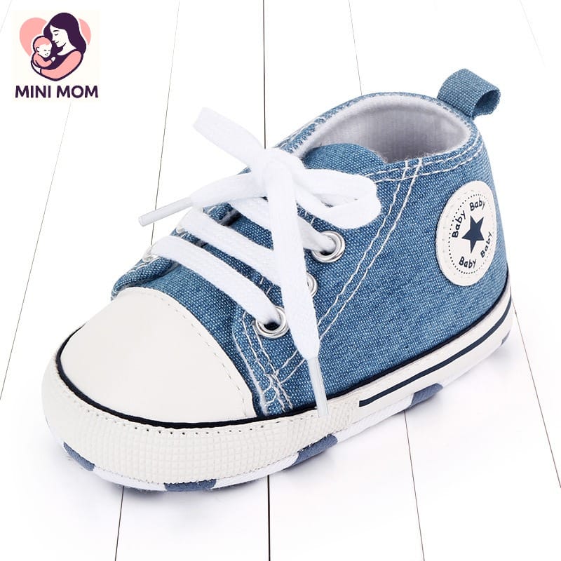 Blue baby sneaker with white laces on a light wooden floor, featuring the Mini Mom brand.