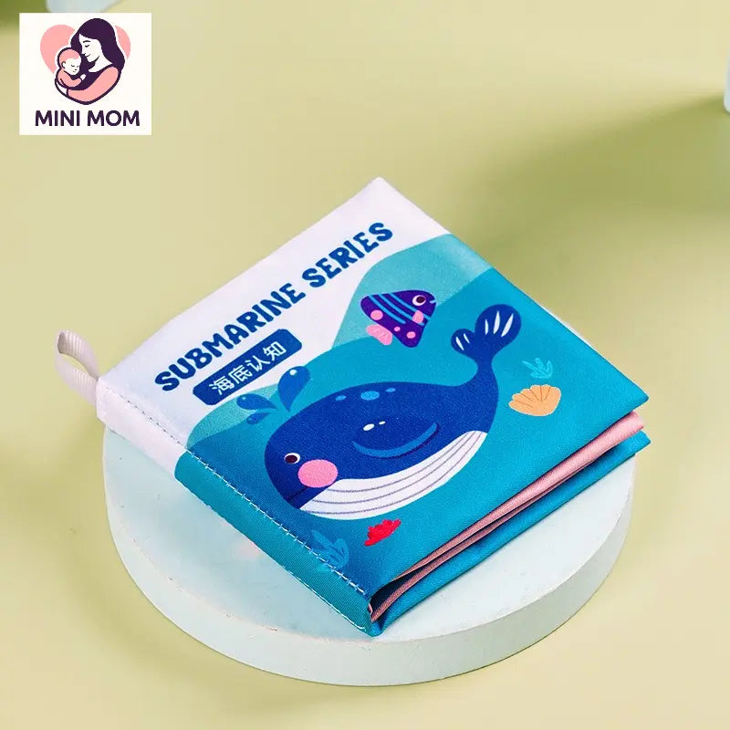 Children's cloth book with whale illustration on a round white platform, featuring the brand 'MINI MOM'.