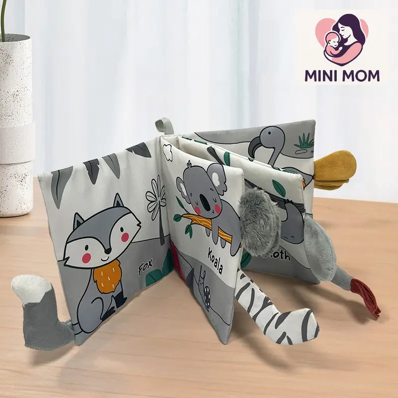 Children's soft book with animal illustrations on a wooden surface, featuring the brand 'MINI MOM'.