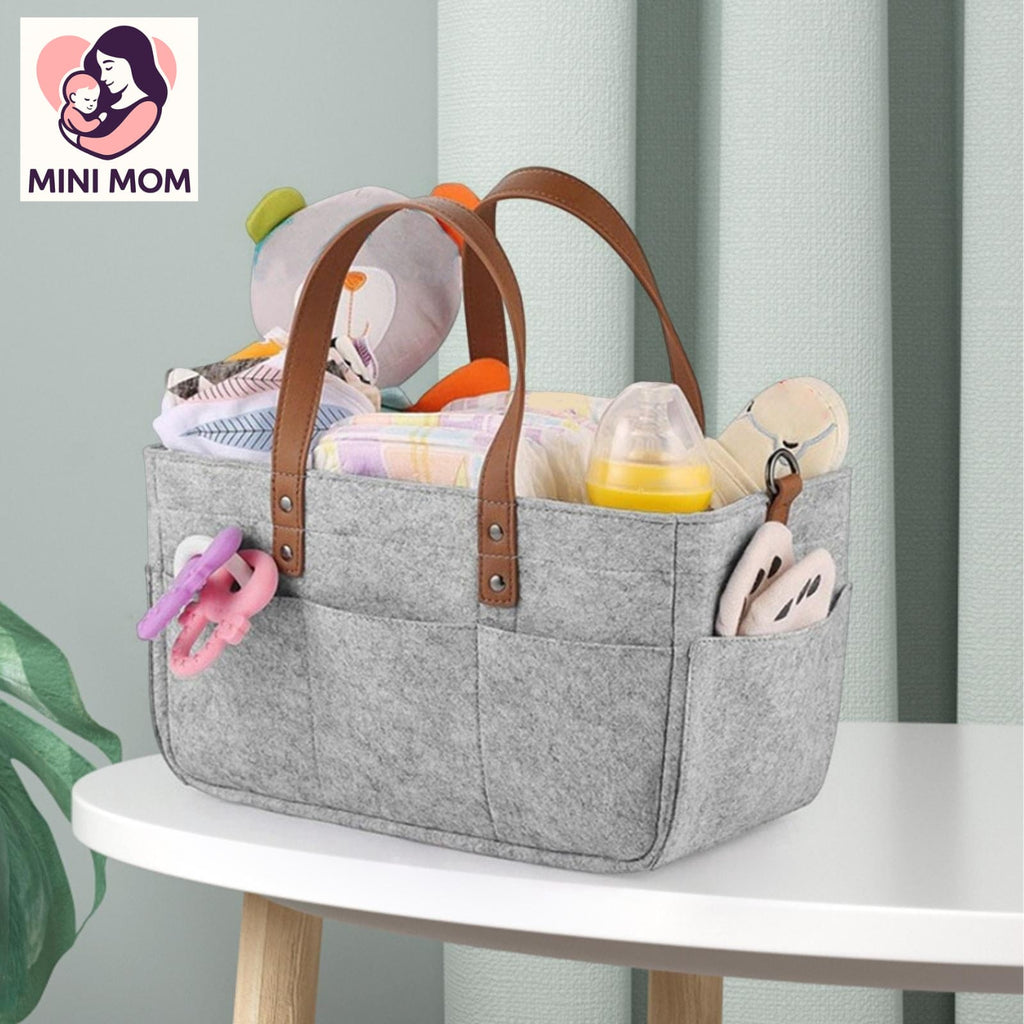 Gray diaper bag with brown handles filled with baby items on a white table, featuring the MINI MOM logo.