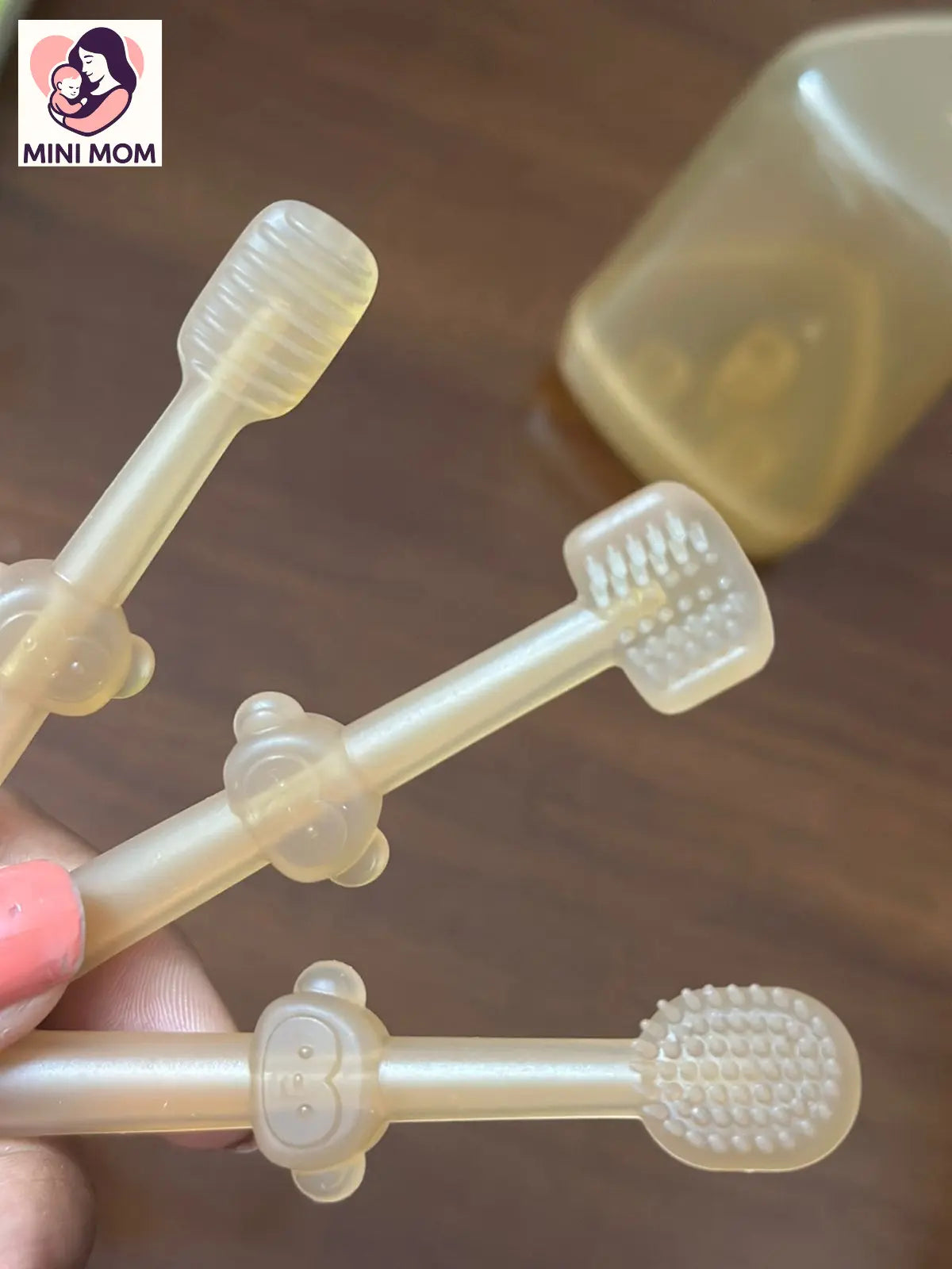 Transparent baby silicone toothbrush held by a hand on a wooden surface with 'MINI MOM' branding.