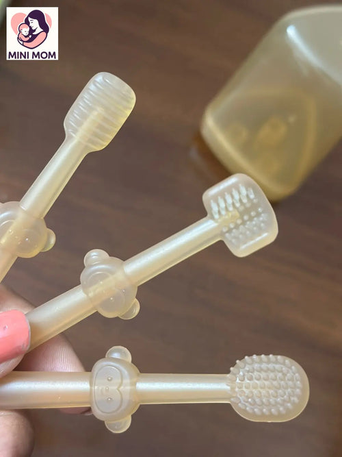 Transparent baby silicone toothbrush held by a hand on a wooden surface with 'MINI MOM' branding.